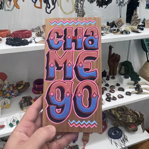 Placa de madeira com pintura colorida escrita "CHAMEGO" em letras grandes e estilizadas, segurada por uma mão em uma loja de artesanato