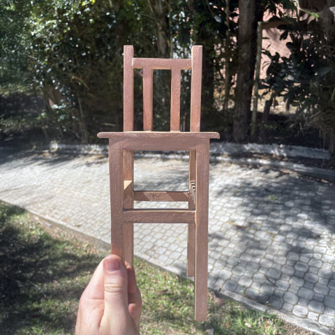 Escultura em madeira de uma pequena cadeira de parede, segurada por uma mão, com fundo de jardim e calçada de pedras