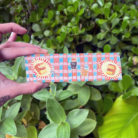 Pintura em madeira com ilustrações de sobremesas e bebidas, segurada por uma mão, em um ambiente verde.