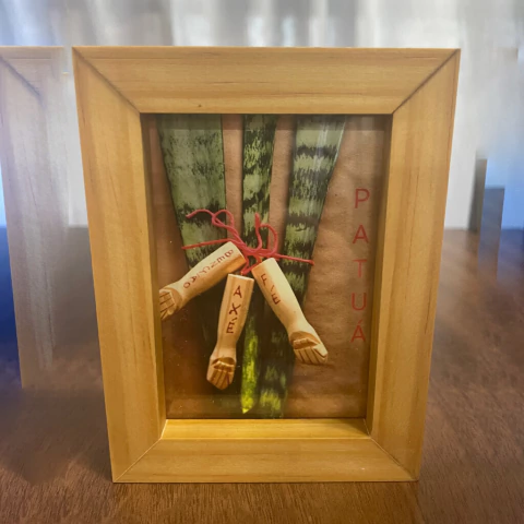 Quadro decorativo com moldura de madeira contendo folhas verdes e três miniaturas de braços com palavras escritas, amarrados por um cordão vermelho