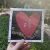 Quadro com coração vermelho e a palavra "Fé" escrito em letras brancas, acompanhado da frase "O amor é assim".