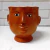 Vaso em cerâmica com formato de cabeça, pintado em laranja com detalhes faciais em preto e branco.