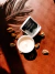Vela aromática de chocolate branco em recipiente branco com tampa preta e decorada, cercada por sementes sobre superfície marrom com sombra de folha