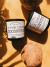 Dois copos pretos com velas aromáticas de coco da Gringo Collection ao lado de um coco seco sobre superfície marrom iluminada por luz natural.
