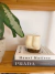Vela decorativa em formato de bumbum sobre uma pilha de livros, com uma planta ao fundo.
