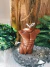 Vela decorativa marrom em formato de torso masculino muscular, posicionada sobre base de pedra cinza com plantas ao fundo