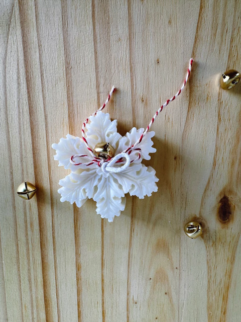 Vela decorativa branca em formato de floco de neve com cordão vermelho e branco e sinos dourados sobre superfície de madeira