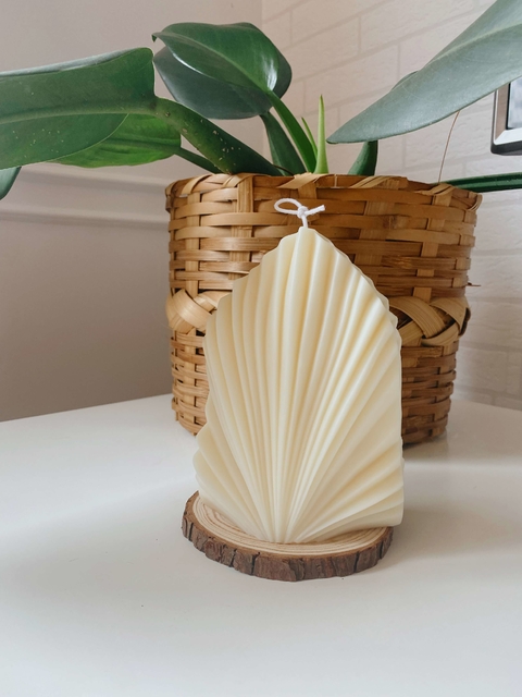 Vela decorativa branca em formato de folha de palma, apoiada em base de madeira, com vaso de planta ao fundo