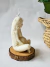 Vela decorativa em forma de figura feminina sentada, com detalhes em relevo e cabelo ondulado, sobre uma base de madeira.