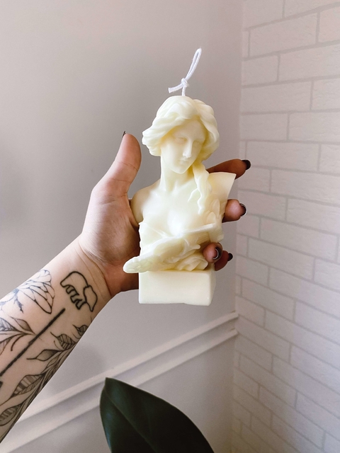 Vela decorativa branca em forma de busto feminino segurando uma harpa, sendo segurada por uma mão com tatuagens