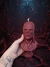 Vela em formato de busto do Vecna detalhado, cor vermelha, segurada por uma mão com tatuagens, em cenário sombrio e místico com raízes e fumaça vermelha.
