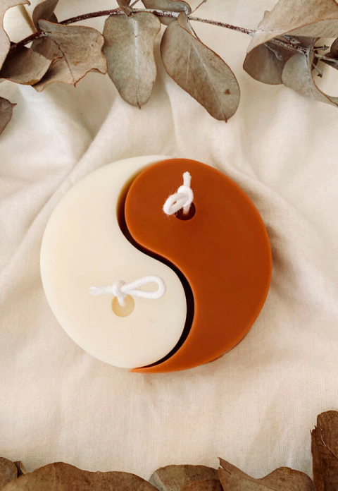 Vela decorativa em formato Yin-Yang com cores off white e marrom sobre tecido claro, cercada por folhas secas