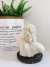 Vela Zeus em formato de busto com detalhes de cabelos ondulados, sobre uma base escura, ao lado de uma planta e um saco de papel decorativo.