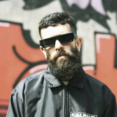 Homem usando óculos de sol Double X, com barba e cabelo curto, em frente a um mural colorido.