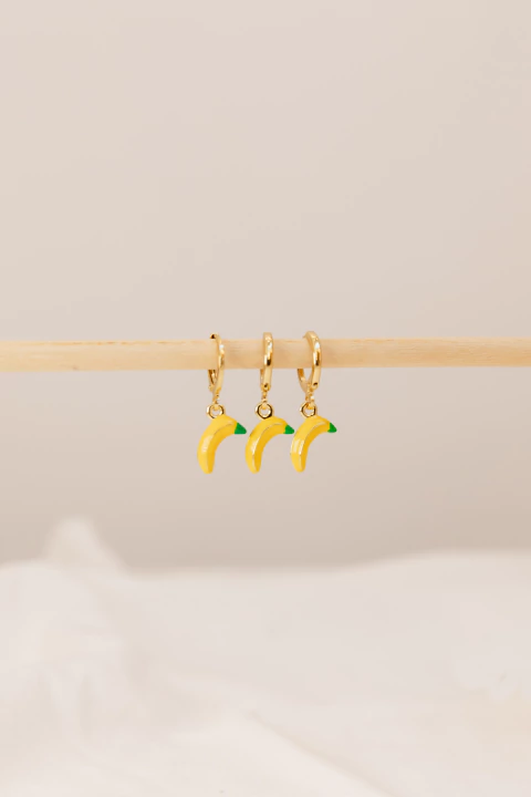 Aros colgantes dorados con charms de banana amarilla y punta verde, colgados de una barra de madera clara.