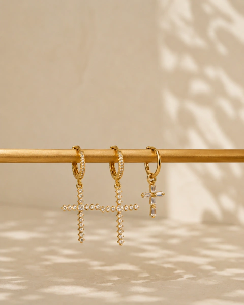 Set de tres aros dorados de Golfield colgados en barra, dos con cruces de strass y uno con cruz lisa, fondo beige con sombras suaves.