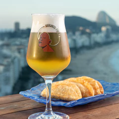 Taça Sommelier Mulher Preta cheia de cerveja com espuma, sobre mesa de madeira, ao lado de prato azul com pastéis, com vista panorâmica da praia ao fundo