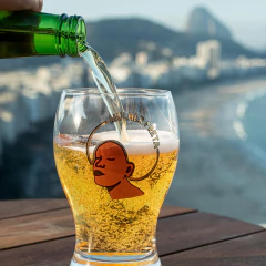 Taça Boston Mulher Preta sendo preenchida com cerveja, com ilustração de rosto feminino negro e texto "Mulher Preta" na taça, ao ar livre com vista para praia e montanha ao fundo