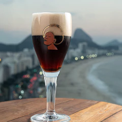 Taça Harzer Mulher Preta com cerveja escura e espuma, sobre mesa de madeira com vista panorâmica da praia e montanhas ao fundo
