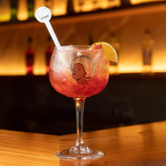 Taça de gin com design Mulher Preta, contendo bebida vermelha com gelo, fatia de laranja e mexedor branco sobre balcão de madeira em bar iluminado