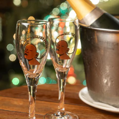 Dupla de taças de champagne transparentes com desenho de rosto estilizado de mulher preta e texto "Mulher Preta" em destaque, ao lado de balde de gelo com garrafa de champagne