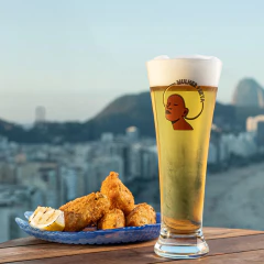Taça Pilsner Mulher Preta cheia de cerveja com espuma ao lado de petiscos em um prato azul, com vista panorâmica da cidade ao fundo