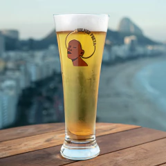 Taça Pilsner com cerveja dourada e espuma, decorada com ilustração de mulher preta e o texto "Mulher Preta", sobre uma mesa de madeira com vista para a praia ao fundo
