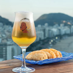 Taça Sommelier Mulher Preta com cerveja dourada e espuma, ao lado de pastel em prato azul sobre mesa de madeira com vista panorâmica da cidade ao fundo