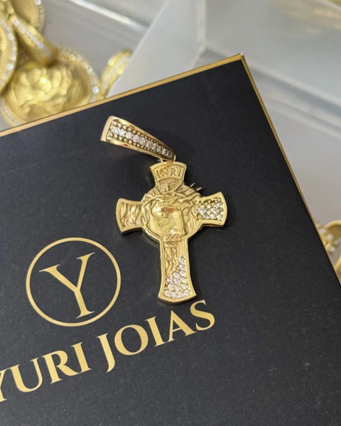 Pingente dourado em forma de cruz com face de Cristo cravejada com pequenas pedras brilhantes