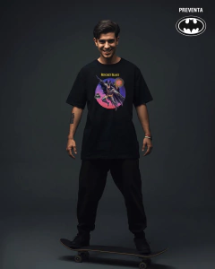 Hombre joven sonriendo con camiseta negra "Rocket Blast" con diseño de Batman y texto amarillo, de pie sobre una patineta