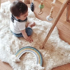 Un bebé sentado sobre un corderito de cuero de oveja color natural, con un arco de colores en el frente y juguetes colgantes alrededor.