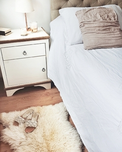 Habitación decorada con una cama blanca, almohada y un corderito de cuero de oveja en el suelo, junto a un velador con velas.