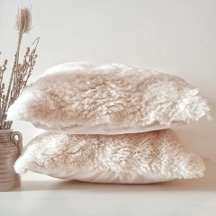 Dos almohadones de cuero de oveja con pelo en color blanco, apilados sobre una mesa, junto a una jarra con flores secas.