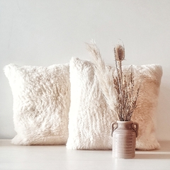 Almohadones de cuero de oveja con pelo en color blanco, acompañados de un jarrón con flores secas sobre una mesa clara.