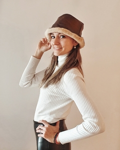 Modelo luciendo un sombrero de cuero marrón con borde de pelaje, vestida con un suéter blanco y pantalones oscuros.