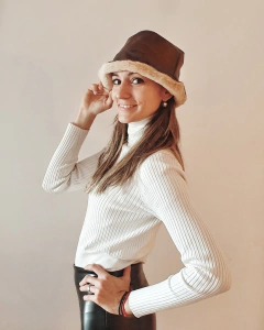 Modelo luciendo un sombrero de cuero marrón con borde de pelaje, vestida con un suéter blanco y pantalones oscuros.