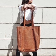 Tote de cuero de vaca color marrón sostenido por una mujer, con fondo de pared blanca.