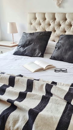 Pie de cama tejido con rayas negras sobre una cama blanca, con almohadas oscuras y un libro abierto.