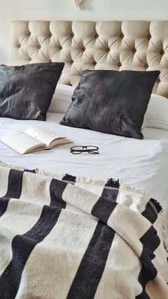 Pie de cama tejido con rayas negras, almohadas oscuras y un libro abierto sobre una cama blanca.