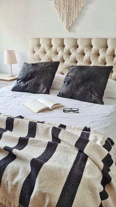 Pie de cama tejido con rayas negras, almohadas negras y un libro abierto sobre una cama blanca.