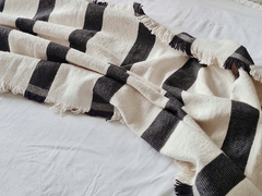 Pie de cama tejido con rayas negras y blancas, con flecos en los bordes, sobre una cama blanca.