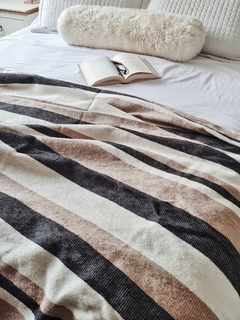 Manta de algodón con rayas marrones y negras sobre una cama, junto a un libro abierto y un almohadón peludo.