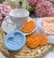 Molde de silicone em formato de biscoito palmier, com biscoitos prontos ao lado de uma xícara branca sobre uma toalha de mesa.