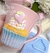 Uma xícara rosa decorada com um cupcake colorido, com glacê e confetes, ao lado de um molde de silicone para cupcakes.