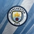 Emblema da camisa Manchester City 2025/2026 em azul celeste e branca, apresentando o logotipo do clube.