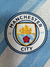 Logotipo do Manchester City em uma camisa azul celeste e branca, destacando um navio e uma rosa.