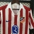 Camisa branca e vermelha do Atlético de Madrid, modelo 25/26, com detalhes em azul e logo da Nike.