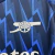Detalhe do escudo bordado do Arsenal em camisa azul marinho e azul com padrão geométrico