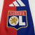 Detalhe do escudo bordado do Olympique Lyonnais na camisa oficial 2025/2026 com listras vermelha e azul