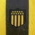 Detalhe do escudo da camisa Peñarol I 2025/2026, com listras amarelas e pretas.